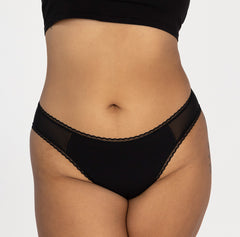 Load image into Gallery viewer, model facing front wearing la coochie organic cotton mid rise brief underwear in midnight black featuring mesh panels and high cut leg
