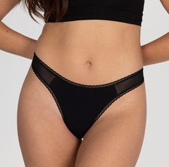 Load image into Gallery viewer, Model showing front view of organic cotton mid rise thong underwear in midnight black, featuring close up of lace detail and mesh panels
