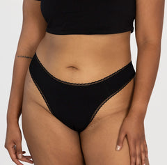 Load image into Gallery viewer, model wearing midnight black la coochie organic cotton mid rise thong underwear, facing front to show off all cotton fabric and flattering contour waist and leg
