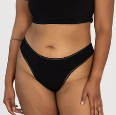 Load image into Gallery viewer, model wearing midnight black la coochie organic cotton mid rise thong underwear, facing front to show off all cotton fabric and flattering contour waist and leg