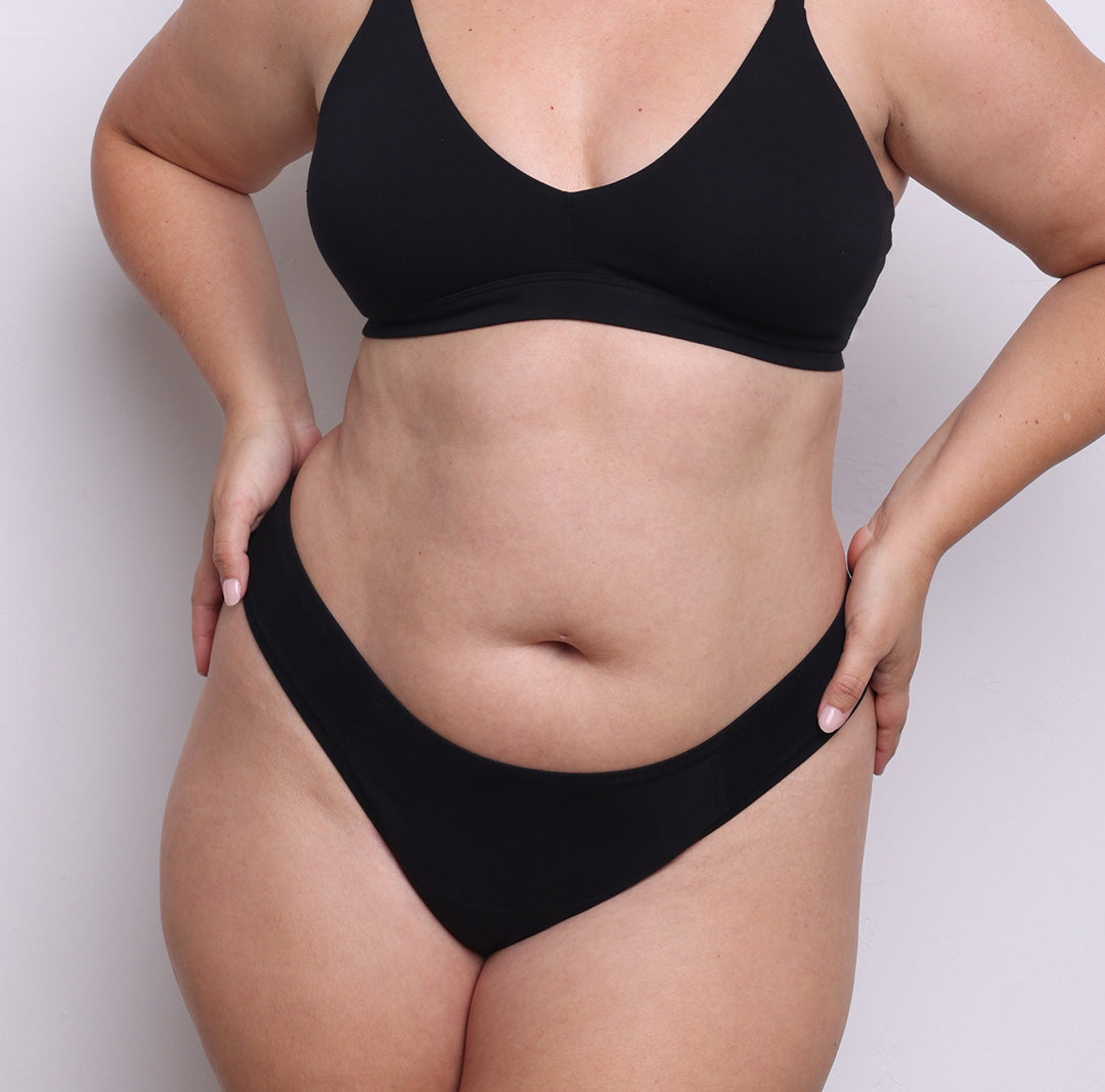 Size L model showing front view of organic cotton dipped front thong in midnight black. Dipped mid rise, high-cut leg and never-tight enclosed elastic