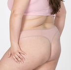 XXL model facing back to show organic cotton mid rise thong underwear in blush pink, features curved thong back to flatter and complement curves, lace detail and mesh panels