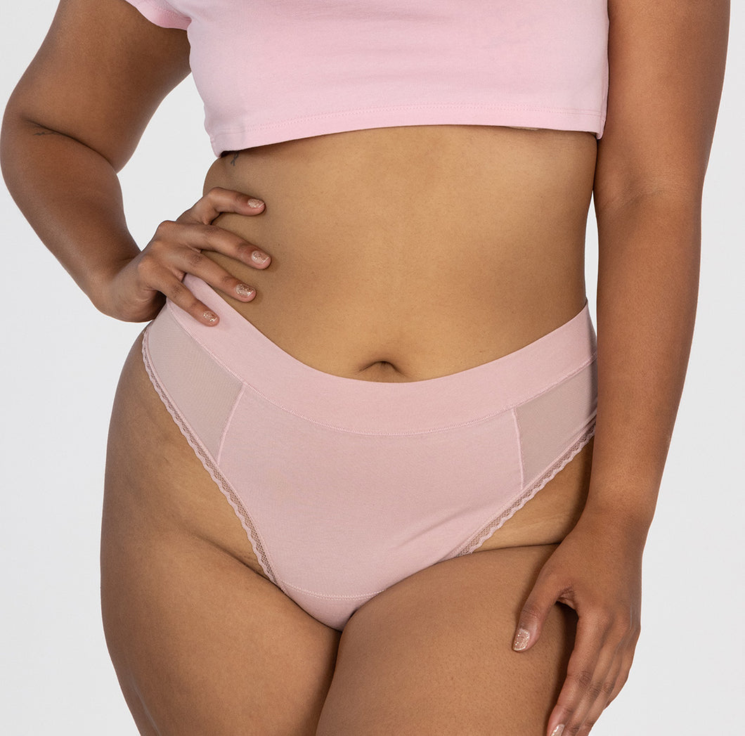 Woman with hand on her hip wearing la coochie organic cotton high waisted thong in blush pink color, featuring lace detail on leg and mesh panels